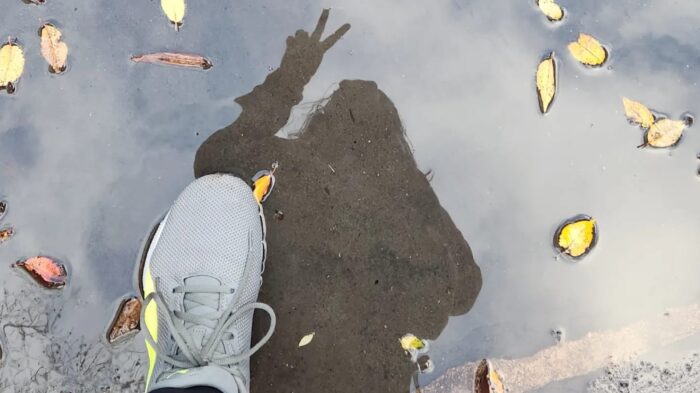 A foot steps into a puddle. In the water’s reflection, the person is visible making a two-finger peace sign. The image symbolizes how accessibility goes beyond physical aspects, reflecting broader, less visible dimensions.